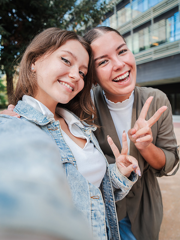 Vertical. Two happy young women smiling and posing for a selfie outdoors, capturing a fun and joyful moment together while enjoying time outside in an urban setting, showing peace sign and friendship